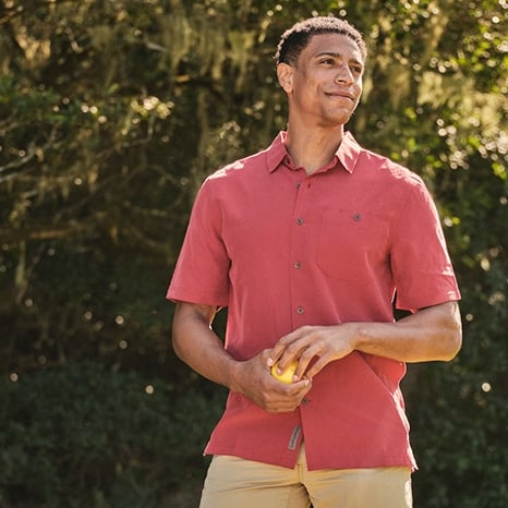 Man holding a ball wearing a red button up shirt and tan shorts smiling.