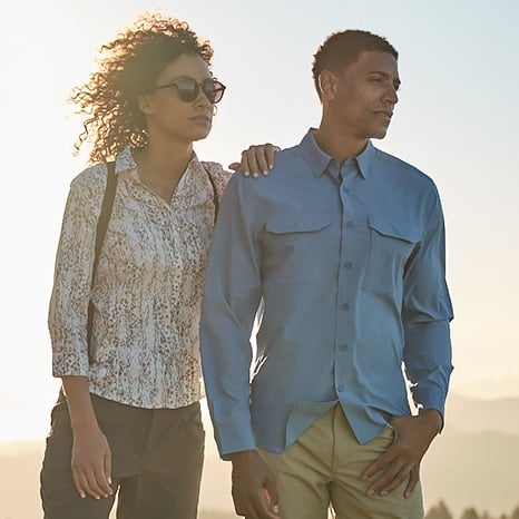 Man in blue shirt with hands in pockets and woman in white patterned shirt leaning on him looking into the distance with hazy and sunny background. 